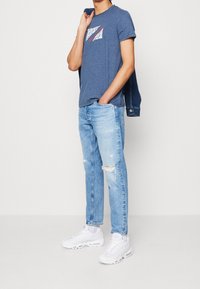 Hombre con camiseta gráfica azul, jeans claros rasgados, zapatillas blancas, sosteniendo una chaqueta de mezclilla sobre su hombro frente a un fondo liso.