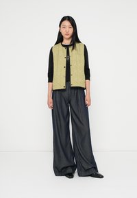 Prošívaná olivově zelená vesta přes černý top s dlouhým rukávem, kombinovaná s volnými tmavými džíny. Obsahuje patentky a volný střih.