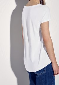 Camiseta blanca de manga corta con un escote redondeado, textura suave y un dobladillo ligeramente curvado, combinada con jeans de mezclilla azules.