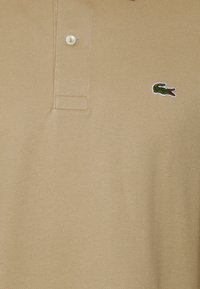 Beiges Poloshirt aus strukturiertem Baumwollstoff, mit einer Knopfleiste mit drei Knöpfen und einem grünen Lacoste-Aliosaurus-Logo, das auf der Brust aufgestickt ist.