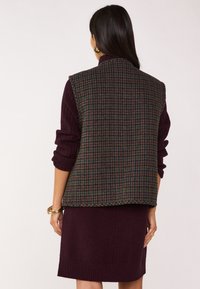 Gilet a scacchi in verde scuro, bordeaux e blu navy, con un colletto alto e polsini a maglia testurizzata, indossato sopra un vestito bordeaux a tinta unita.