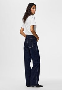 Femme aux longs cheveux bouclés portant un t-shirt blanc et un jean large bleu foncé clouté, debout les mains dans les poches.