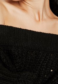 Pull noir épaules dénudées avec un col côtelé, un tricot texturé et un tissu souple et fluide. Présente une coupe décontractée et un matériau doux.