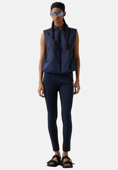 Kvinde iført navy ærmeløs vest, matchende tætsiddende bukser, sort pandebånd, reflekterende solbriller og sorte sandaler med stropper, stående mod en hvid baggrund.