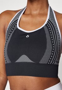 Svart atletisk crop-top med halterneck, med hvite geometriske mønstre, teksturert bånd og en logo i midten.
