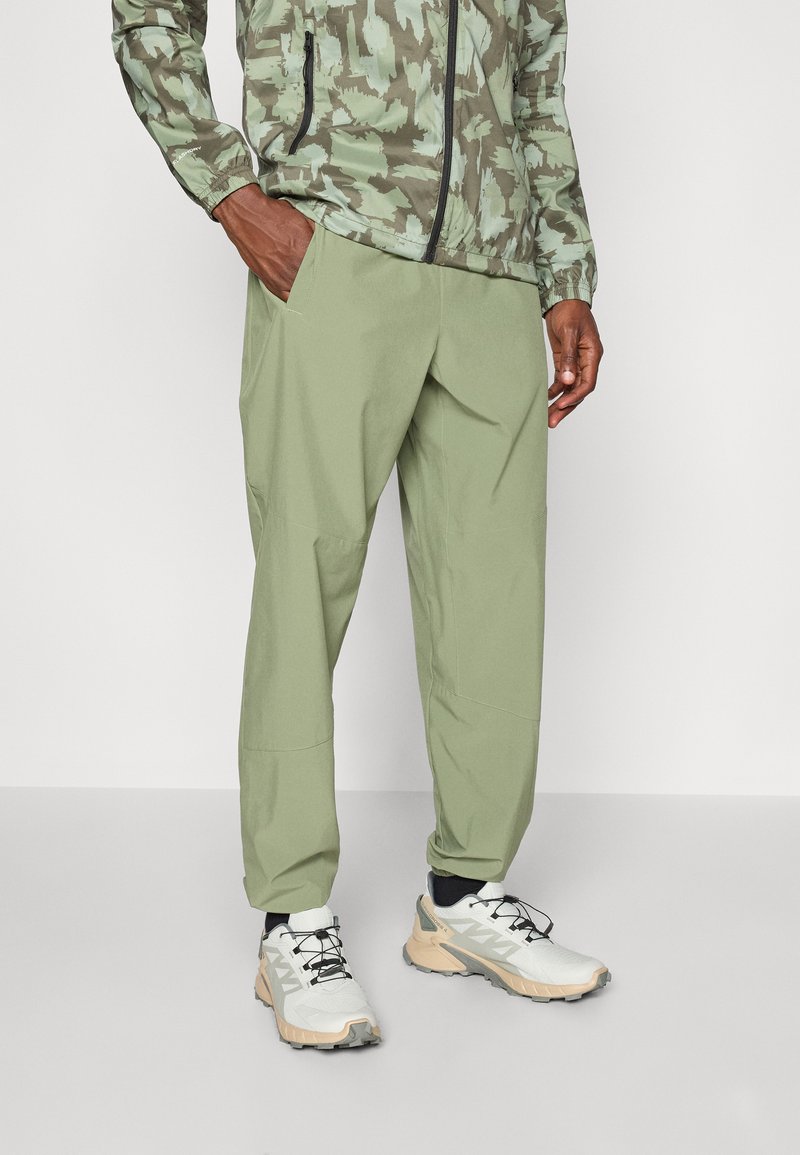 Pantaloni de un verde olive deschis, realizati dintr-un material usor, cu o croiala lejera, buzunare laterale si manșete conice, asortați cu adidași albi și bej.