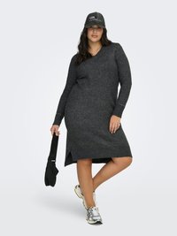 Kvinna klädd i en mörkgrå stickad klänning, keps med texten "Los Angeles", håller en svart väska i ena handen och bär silverfärgade sneakers, står med ena benet korsat.