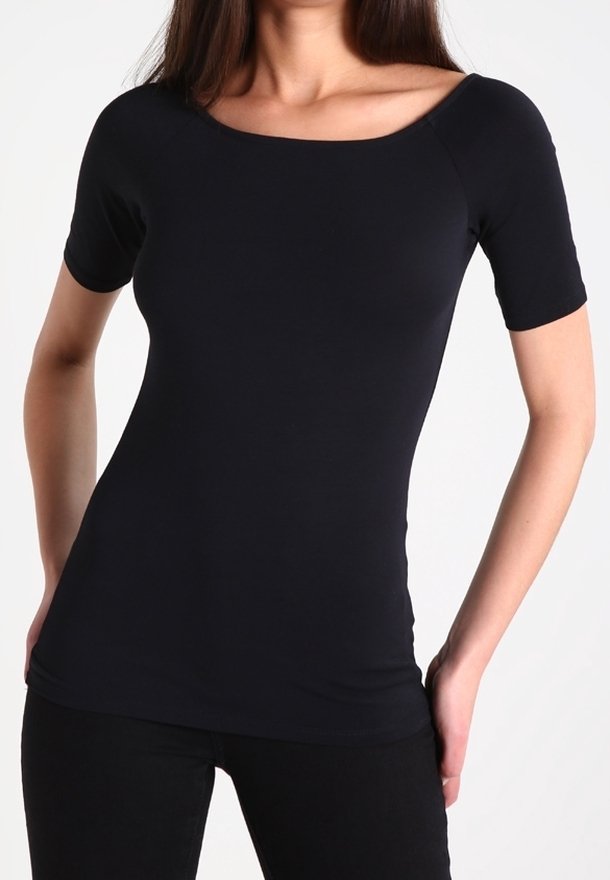 Femme portant un haut noir ajusté à manches courtes avec un large décolleté en arrondi et un pantalon noir, debout les mains dans les poches.