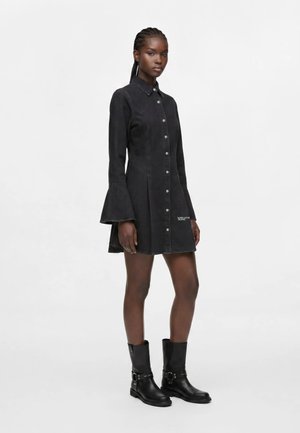 Femme portant une robe noire boutonnée avec des manches évasées et des bottes noires mi-mollet, debout devant un fond blanc uni.