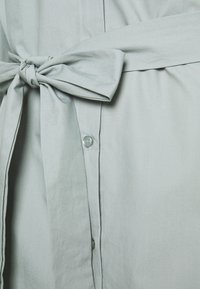 Svetle šedé textilní šaty s knoflíkovou légou a vázaným pasem. Textura je hladká, s jemným leskem a měkkými záhyby.