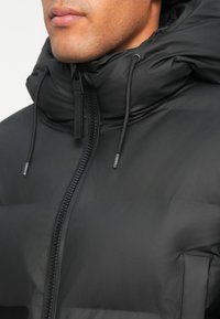 RAINS Cappotto lungo Rains Unisex in tessuto 100% poliestere rivestito in poliuretano, modello Alta Long Puffer Jacket W3T4, senza piume, collo alto con cappuccio regolabile con coulisse, tasche late