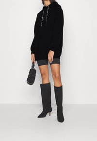 Mujer con una sudadera negra, pantalones cortos de ciclista grises, botas de tacón altas negras hasta la rodilla y sosteniendo un bolso de red negro.