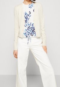 Cardigan crema con una consistenza morbida, cinque bottoni sulla parte anteriore, indossato sopra una maglietta bianca con motivi floreali blu, abbinato a pantaloni bianchi a gamba larga.