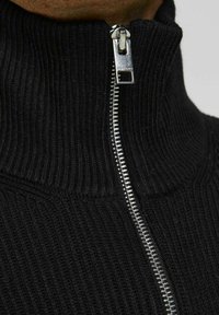 Gros plan sur une personne portant un pull noir en maille côtelée avec une fermeture éclair argentée partiellement remontée jusqu'au cou.