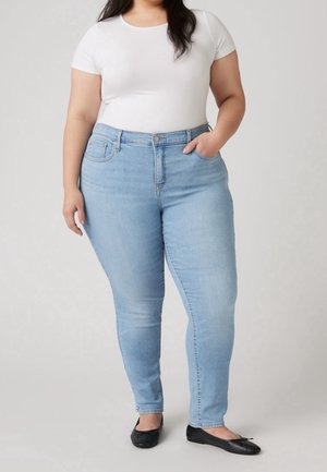 Femme portant un jean skinny taille haute bleu clair, un t-shirt blanc à manches courtes, des ballerines noires, une main dans la poche, sur un fond uni.