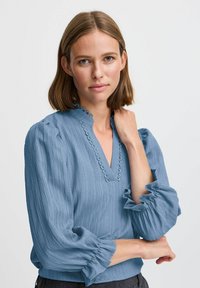 Blusa azzurro chiaro, strutturata, con scollatura a V, maniche arruffate e bordi ondulati lungo il colletto. Tessuto liscio con una vestibilità comoda.