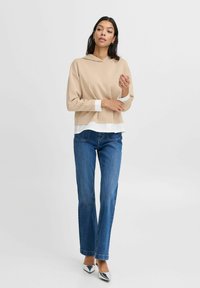 Beige hoodie met lange mouwen, witte manchetten, gelaagd over een witte blouse, gecombineerd met blauwe jeans en zilveren puntige hakken.