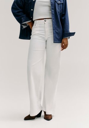 Donna che indossa pantaloni bianchi a vita alta e gamba larga, top corto bianco, giacca di jeans blu scuro e stivali marroni con tacco, in piedi con una mano in tasca.