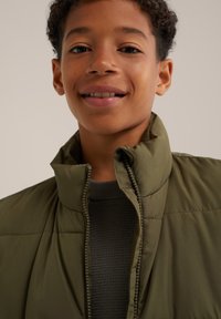 Olivgrön puffjacka med hög krage, som har dragkedjestängning och ett vadderat mönster, båd iklädd över en mörk, texturerad tröja.