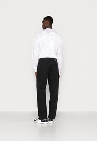 Black denim pants and a white, patterned long-sleeve shirt; shirt features subtle pastel tones and graphic details on the back.