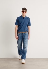 Μπλε πόλο μπλούζα από βαμβάκι, συνδυασμένη με ανοιχτού χρώματος τζιν παντελόνι. Άτομο που φοράει λευκά αθλητικά παπούτσια και καφέ ζώνη, με γυαλιά ηλίου.