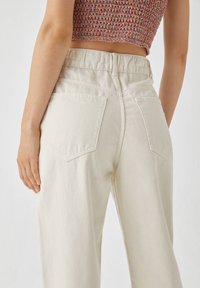 Jeans de tiro alto beige claro con cinturilla elástica, que cuentan con dos bolsillos traseros y un corte relajado. La tela tiene una textura suave.
