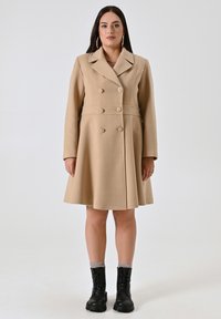 Cappotto in misto lana beige con design svasato, bottoni doppi, colletto a punte e maniche lunghe. Abbinato a stivali da combattimento neri.