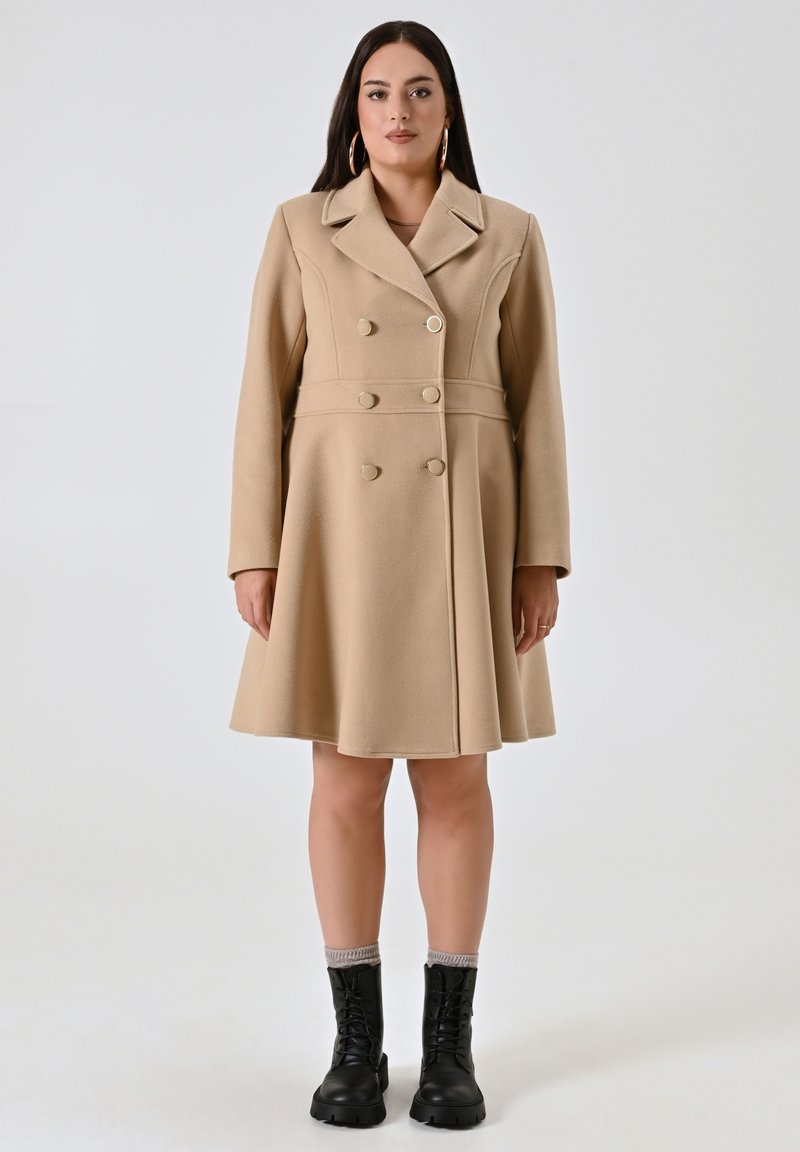 Cappotto in misto lana beige con design svasato, bottoni doppi, colletto a punte e maniche lunghe. Abbinato a stivali da combattimento neri.