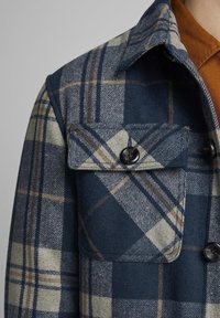 Kostkovaná bunda v námořnické, béžové a hnědé barvě. Vyrobena z texturované tkaniny, obsahuje zapínací klopu a klasický límec.