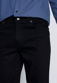 Schwarze Denim-Jeans mit geradem Schnitt, ausgestattet mit silbernen Beschlägen und Fronttaschen. Kombiniert mit einem blauen Hemd mit Knöpfen.