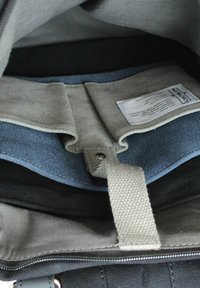 Innenansicht einer Tasche mit grauem Stofffutter, Denim-Akzenten, schwarzen Bereichen und einem Etikett. Beinhaltet einen Riemen und eine Druckknopfschlussdetail.