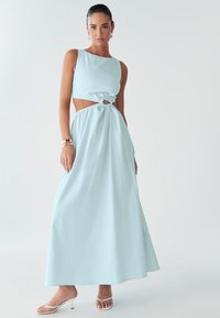 Modèle portant un ensemble deux pièces bleu clair avec un détail découpé, se tenant devant un fond uni, chaussé de sandales blanches à lanières et d'accessoires.