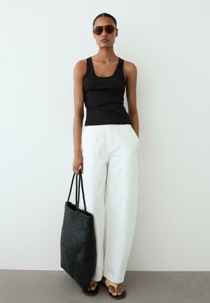 Femme portant un débardeur noir, un pantalon large blanc, des lunettes de soleil, des sandales jaunes, tenant un grand sac cabas noir tissé contre un mur blanc.