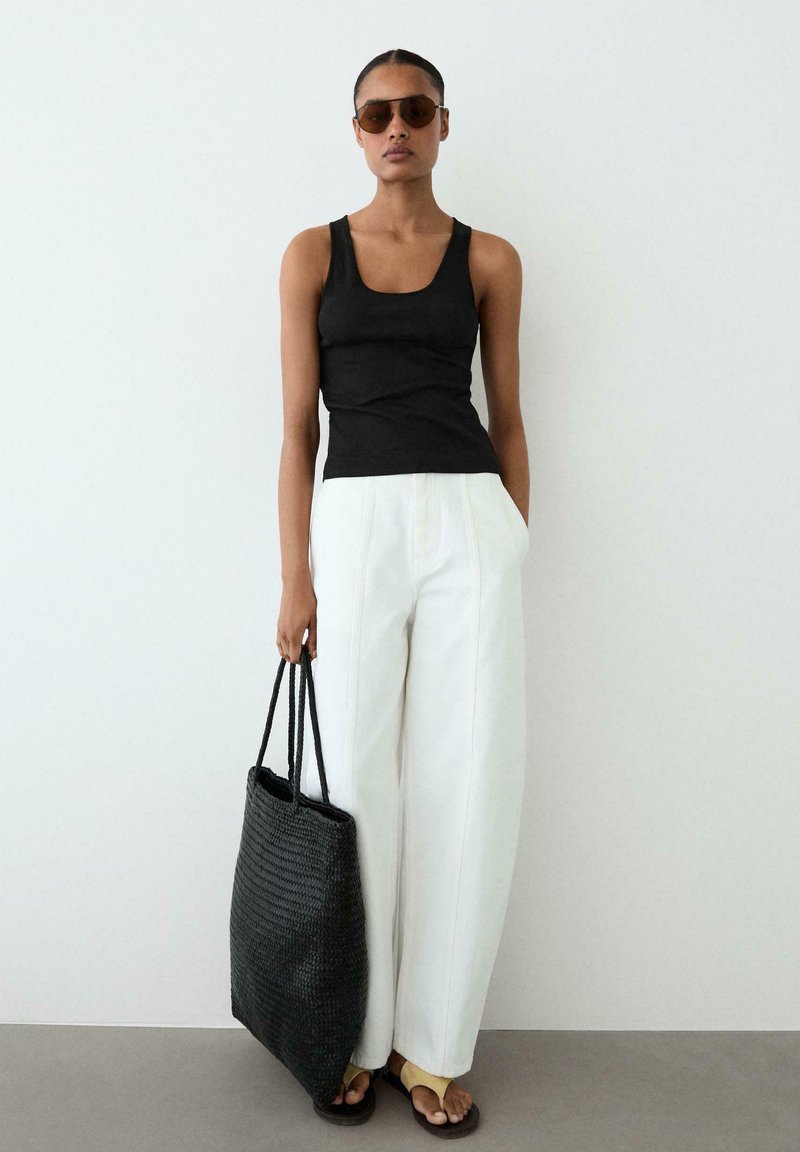 Frau trägt schwarzes Tanktop, weiße weite Hosen, Sonnenbrille, gelbe Sandalen, hält große schwarze gewebte Tote-Bag vor weißer Wand.
