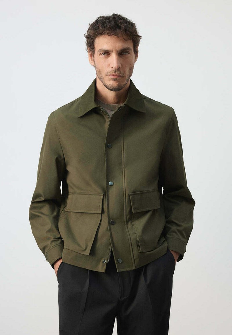 Homme portant une veste verte boutonnée avec des poches à rabat et un pantalon noir, debout devant un fond clair uni.