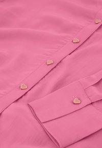 Roze blouse met een gladde stoftextuur, een knoopsluiting versierd met hartvormige knopen met gouden accenten, en lange mouwen met knoopmanchetten.