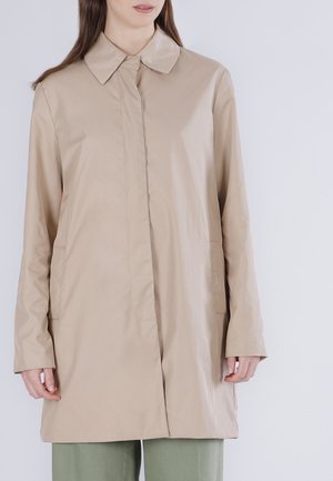 Nainen, joka pukeutunut vaaleanbeigeen pitkähihaiseen takkiin, jossa on kaulus ja edessä kiinnitys, yhdistettynä vaaleanvihreisiin housuihin, seisoo yksivärisen taustan edessä.