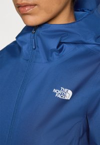 Veste bleue en tissu lisse avec un col haut et une fermeture éclair à l'avant. Le logo The North Face est en blanc sur le haut à gauche.