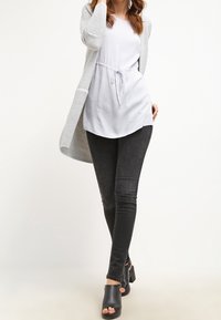 Cardigan grigio lungo con maniche a strisce sopra una blusa bianca, abbinato a jeans skinny neri e sandali chunky con tacco nero.