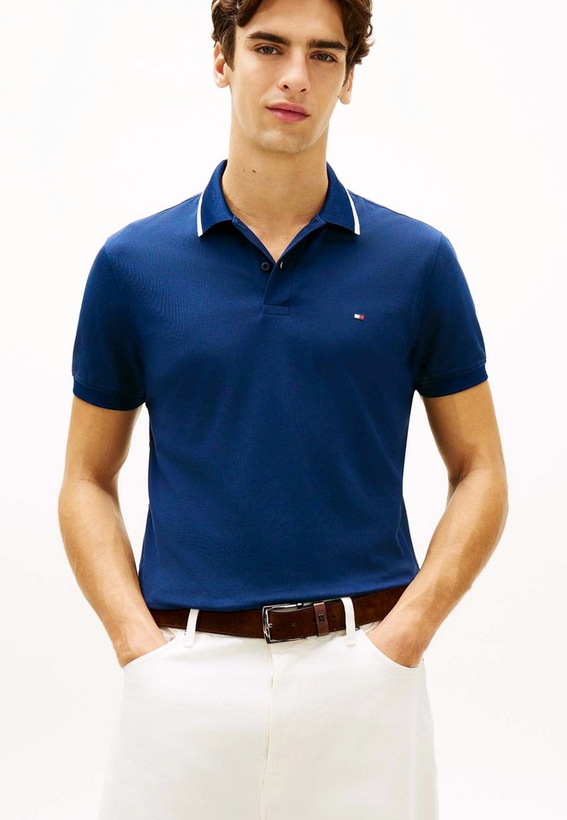 Jeune homme portant un polo bleu marine avec un liseré blanc, un pantalon blanc et une ceinture marron, debout les mains dans les poches.