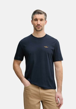 Uomo con capelli corti e grigi che indossa una t-shirt blu navy con un piccolo emblema dorato sul petto e pantaloni beige, in piedi con una mano in tasca.