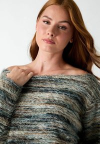 Jeune femme à la peau claire et aux longs cheveux châtain clair porte un pull rayé duveteux à épaules dénudées, tenant l'encolure d'une main.