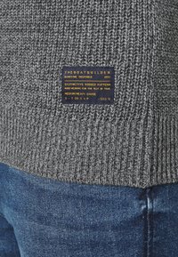 Grå ribbad stickad tröja med en texturerad tyg och en marinblå etikett med guldtext som anger varumärke och skötselråd.