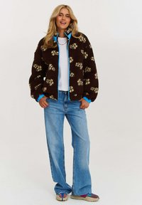 Hnědá fleecová bunda se žlutými květinovými vzory, modrými zipy a nátepníky. V kombinaci se světle modrými širokými džínami a vícebarevnou obuví.