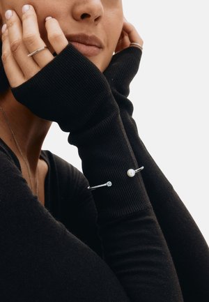 Pulseras de plata con un acento de perla y una piedra preciosa, llevadas en una muñeca cubierta por una manga larga negra acanalada, mostrando una vista de cerca.