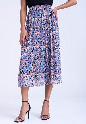Donna che indossa una gonna midi floreale colorata con vita arricciata e sandali neri con tacco alto, in piedi su uno sfondo chiaro uniforme.