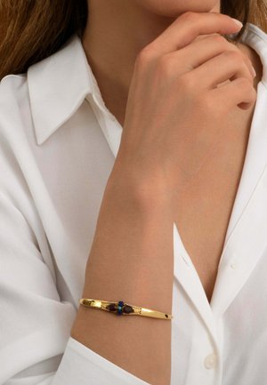 Femme portant une chemise blanche touche son visage avec sa main gauche, mettant en valeur un bracelet en or avec des pierres précieuses rouges et bleues à son poignet.