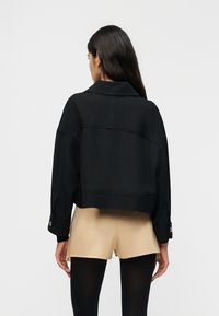 Γυναίκα με μακριά σκούρα μαλλιά, φαίνεται από πίσω, φοράει μαύρο cropped σακάκι, μπεζ σορτς και μαύρα καλσόν σε απλό φόντο.