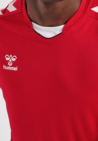 Červený sportovní dres z měkké, lehké tkaniny. Má véčkový výstřih, černé lemování a bílé logo s vzorem na ramenech.