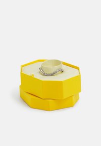 Gelbe sechseckige Schmuckschatulle mit strukturiertem Oberflächen. Darin liegt ein silbernes Armband mit gewelltem Design und funkeltenden Akzenten.